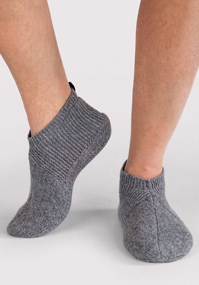 A person stands on a light surface wearing Woolx Mens Gripper Slippers in Gentle Grey. These thick, knitted slippers feature a textured pattern and cover the ankles.
