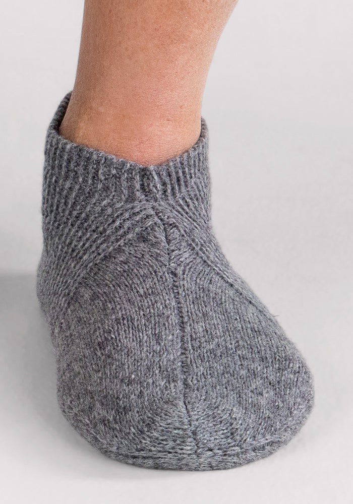 A close-up of a person's foot wearing Woolx Mens Gripper Slippers in Gentle Grey, crafted from knitted merino wool, set against a light background.