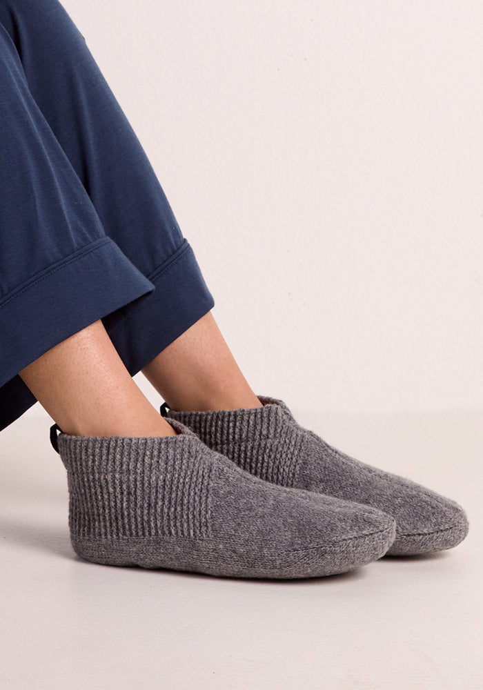 A person sits with crossed feet on a light floor, showcasing the comfort of Woolx Womens Gripper Slippers - Gentle Grey, paired with dark blue pants against a plain background.