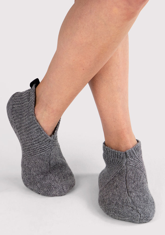 A person with crossed legs is wearing Woolx Womens Gripper Slippers in Gentle Grey—these cozy, textured slipper socks with a small black heel loop offer comfort and stability on any surface.