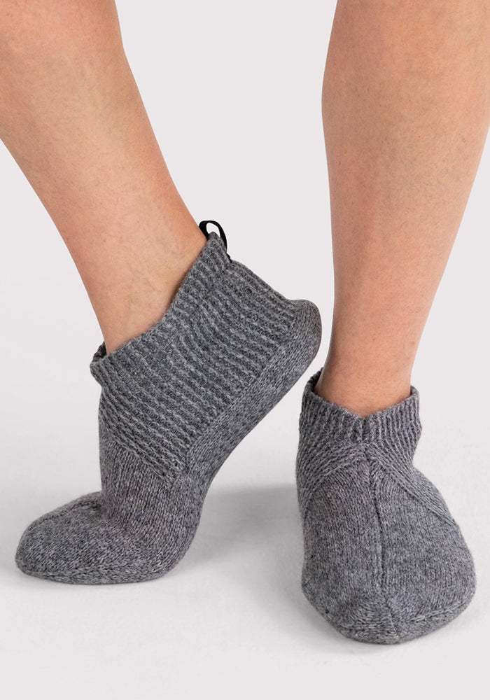 A woman models Woolx Womens Gripper Slippers in Gentle Grey, standing on a light surface with one foot slightly raised—an ideal way to showcase these cozy, non-slip slippers.