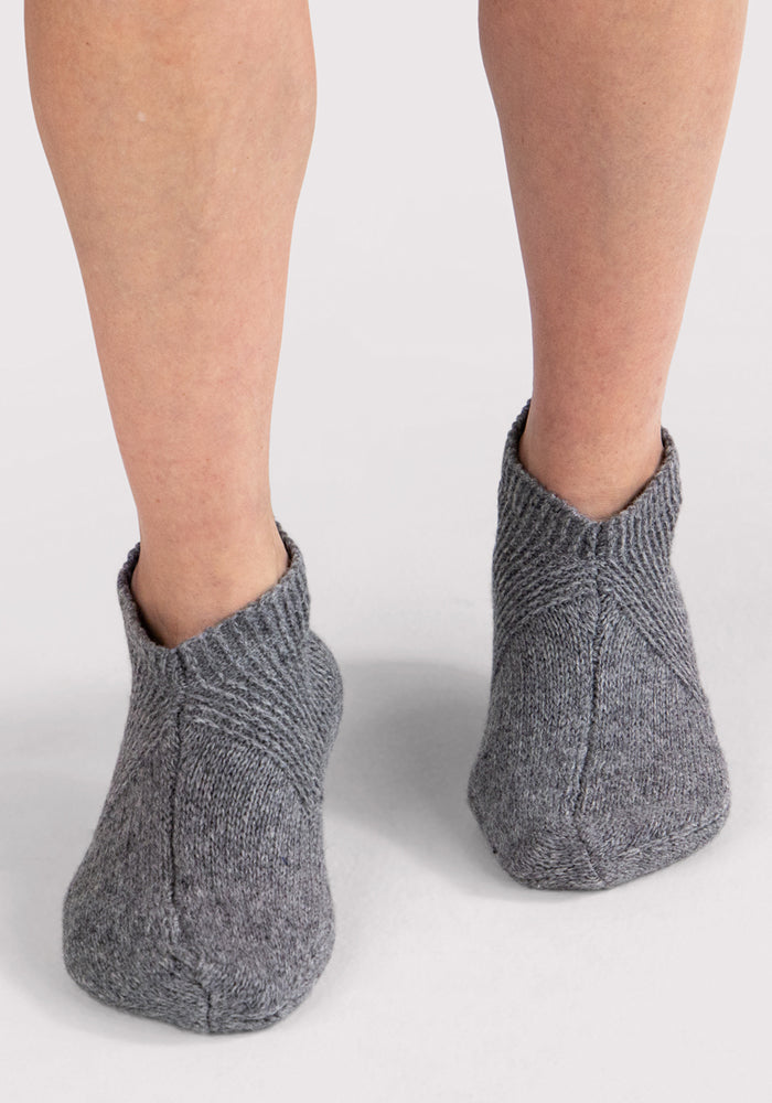 A close-up back view shows two feet in Woolx Womens Gripper Slippers - Gentle Grey, featuring a textured ribbed pattern and ankle coverage on a light background.