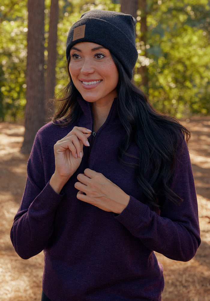 A woman smiles in a wooded area, wearing the Woolx Collins Half Zip Sweater in Storm Cloud and a dark beanie, as she unzips the sweater slightly with sunlight streaming through the trees.