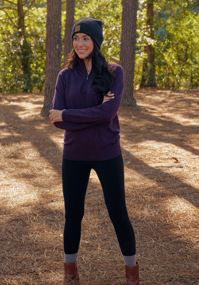 A woman stands outdoors on a pine-covered forest floor, smiling with arms crossed. She wears the Woolx Collins Half Zip Sweater in Deep Plum, a black beanie, black leggings, gray socks, and brown boots among tall trees and green foliage.