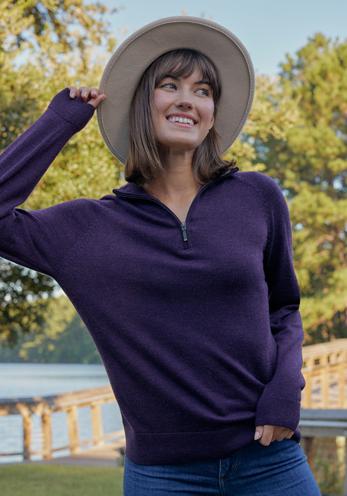 A smiling woman in the Woolx Collins Half Zip Sweater in Wild Salmon and a beige hat stands outdoors near a wooden walkway by the water, with trees in the background.