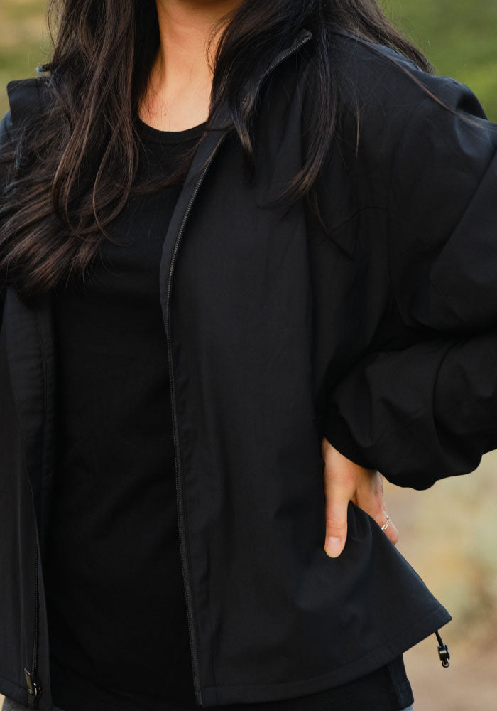 Woman wearing the Woolx Cooper Windbreaker - Black stands outdoors, hand on hip, showcasing zippered pockets.