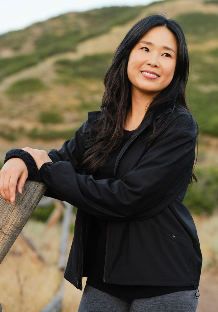 Woman in the Woolx Cooper Windbreaker - Black, with zippered pockets, smiles by grassy hills while leaning on a fence.