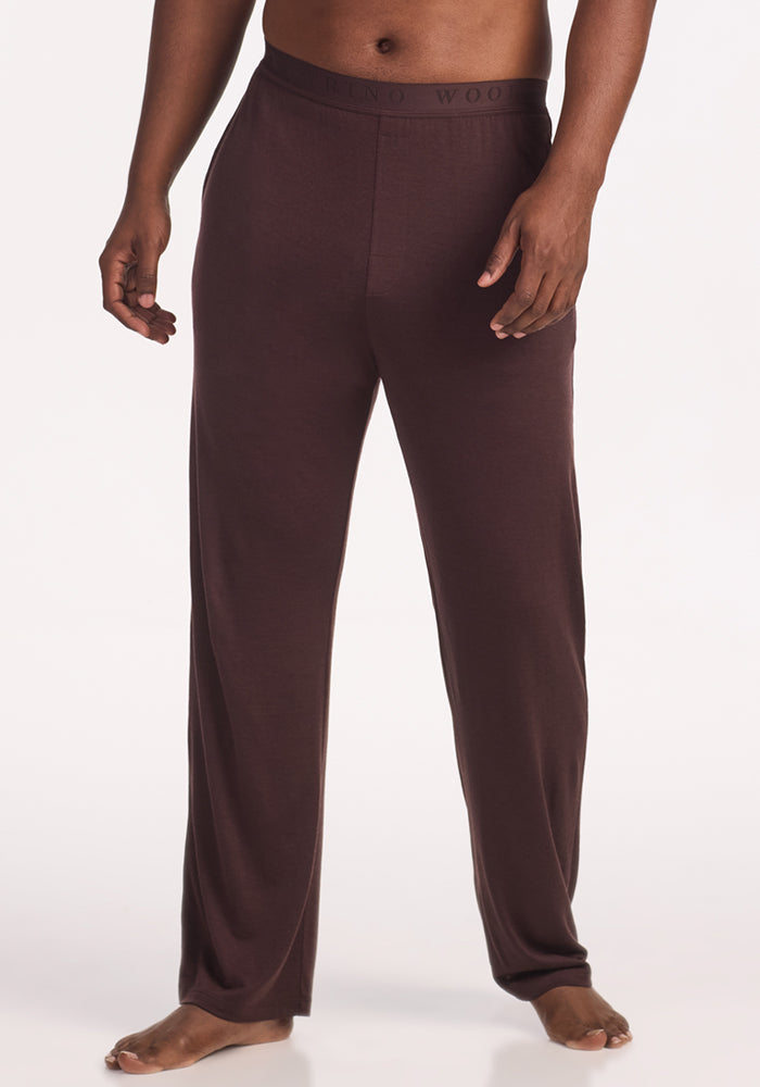A person stands barefoot against a plain white background, wearing Woolx Arlo Lounge Pants in Chocolate Plum—loose-fitting Merino wool pants with an elastic waistband. 
