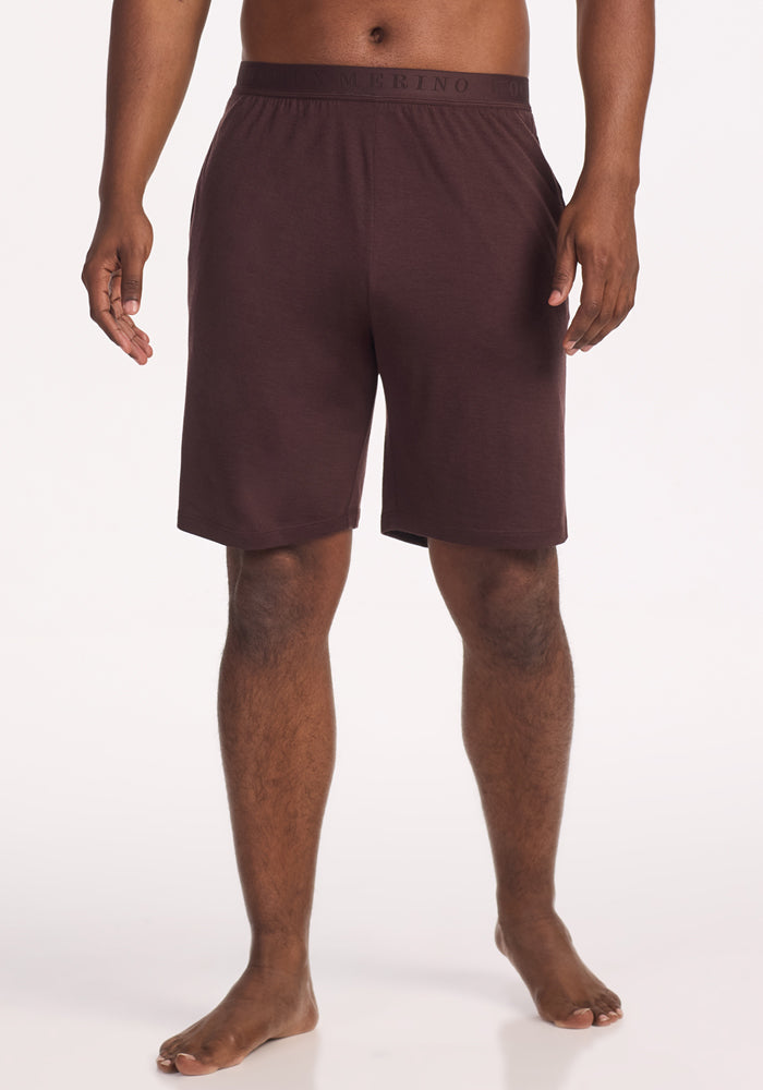 A person stands barefoot against a white background, showing only their lower torso and legs while wearing Woolx Archer Sleep Shorts in Chocolate Plum. The shorts feature an elastic waistband and are made from Merino wool. 
