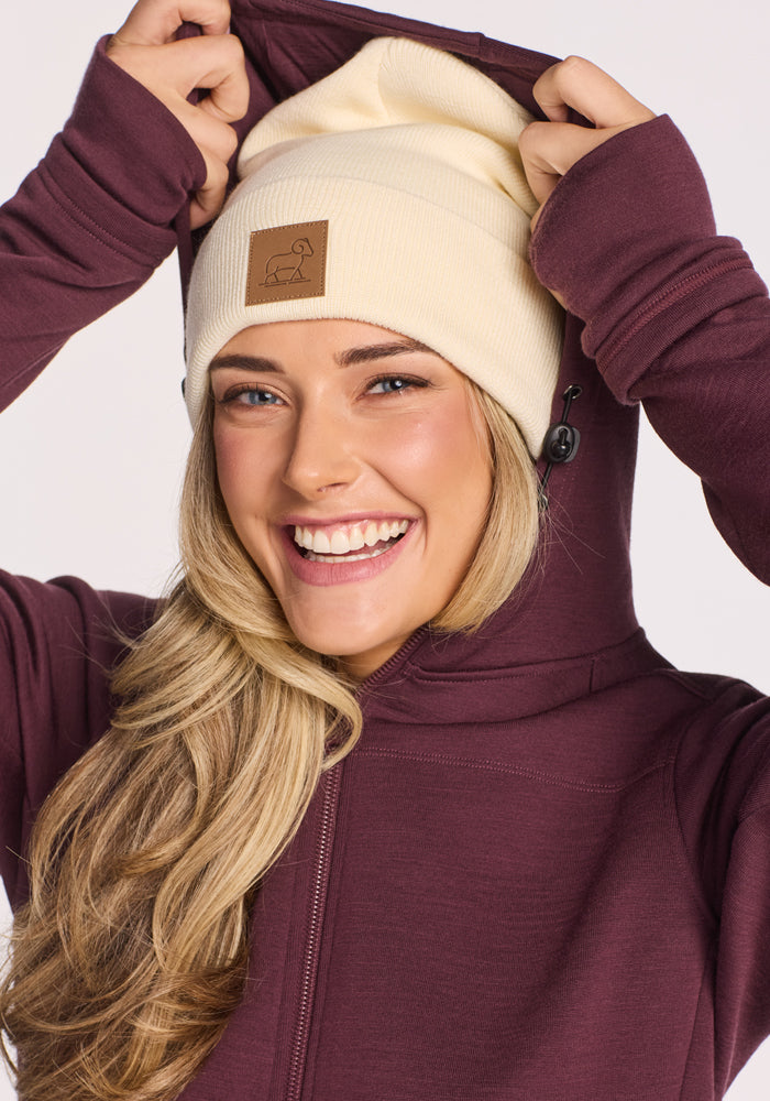 A smiling woman with long blonde hair wears the Woolx Baylor Beanie in Sweet Cream and a maroon zip-up hoodie, holding the hood near her head.