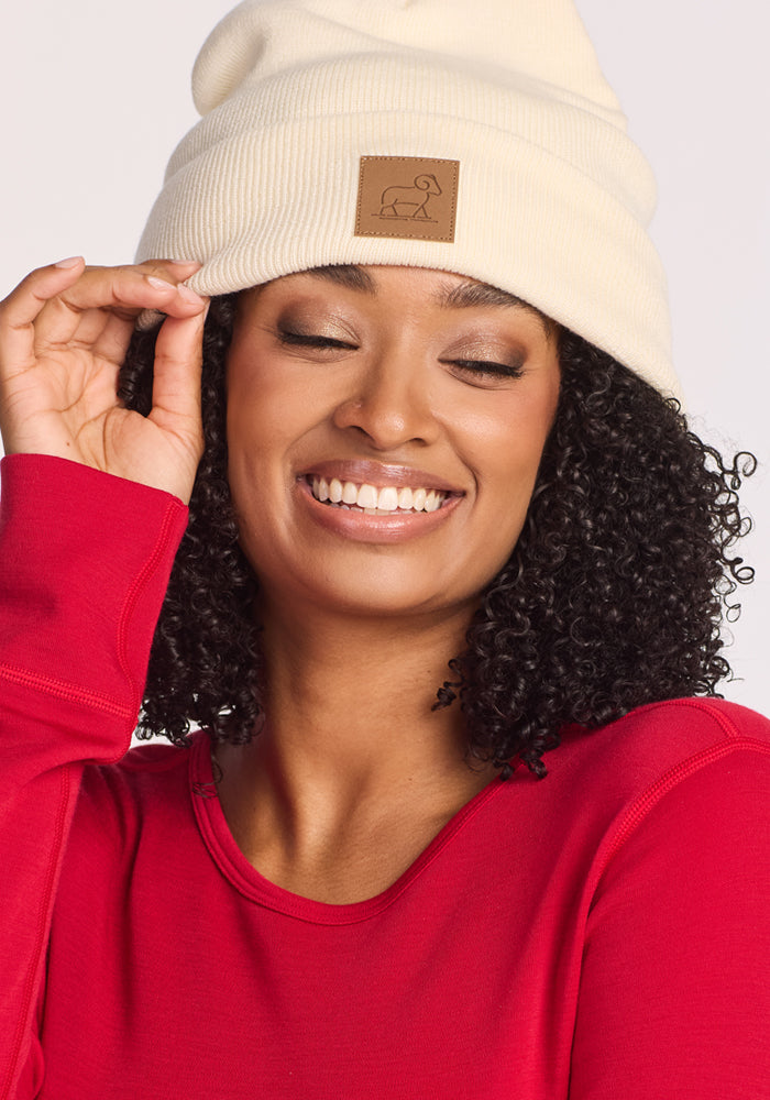A smiling woman with curly hair wears the Woolx Baylor Beanie in Sweet Cream and a red long-sleeve shirt. She touches the edge of her beanie with one hand, eyes closed, looking cheerful.