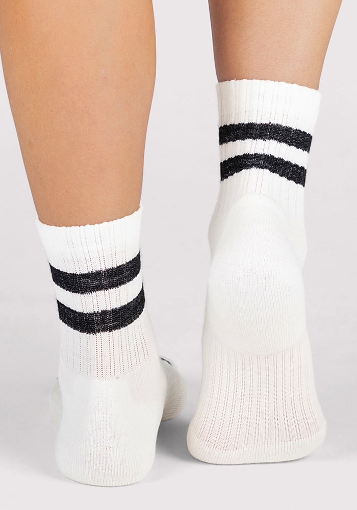 A close-up shows a person wearing Woolx Womens Varsity Mini Crew Socks - Classic Stripe, featuring two black stripes near the top, standing against a plain light background.