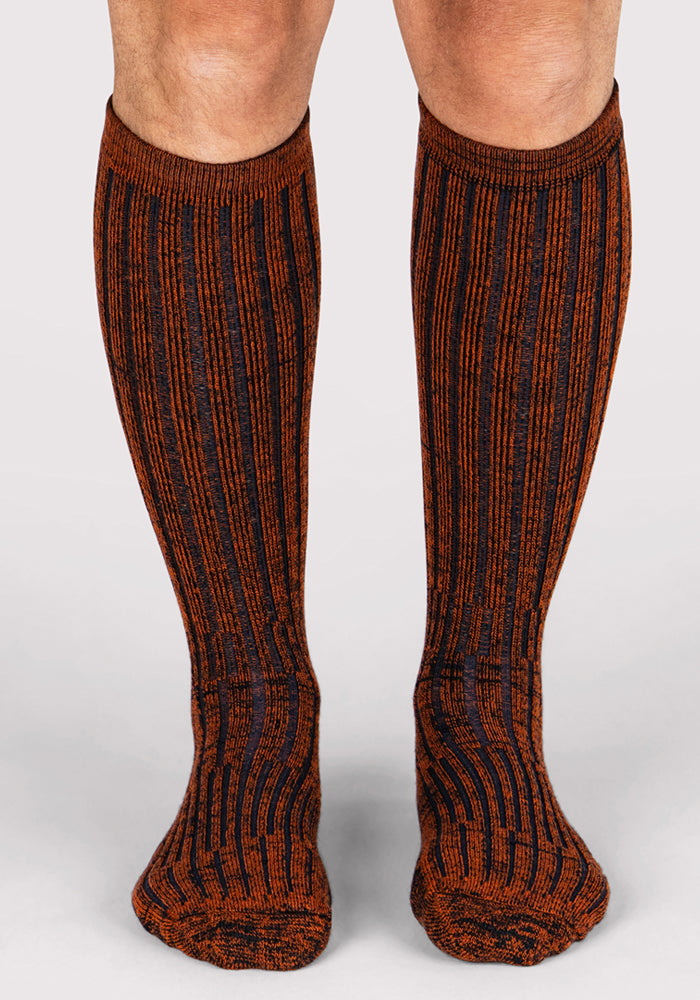 A pair of feet wearing Woolx Mens Joey Mid Calf Socks in Cinnamon Spice, featuring black and orange vertical stripes, is displayed on a light background.