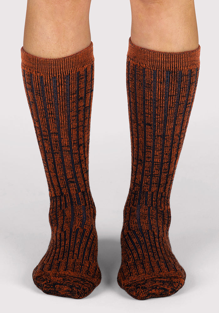 A pair of legs wearing Woolx Womens Joey Mid Calf Socks in Cinnamon Spice, featuring a ribbed marled orange and black pattern, standing on a light gray background.