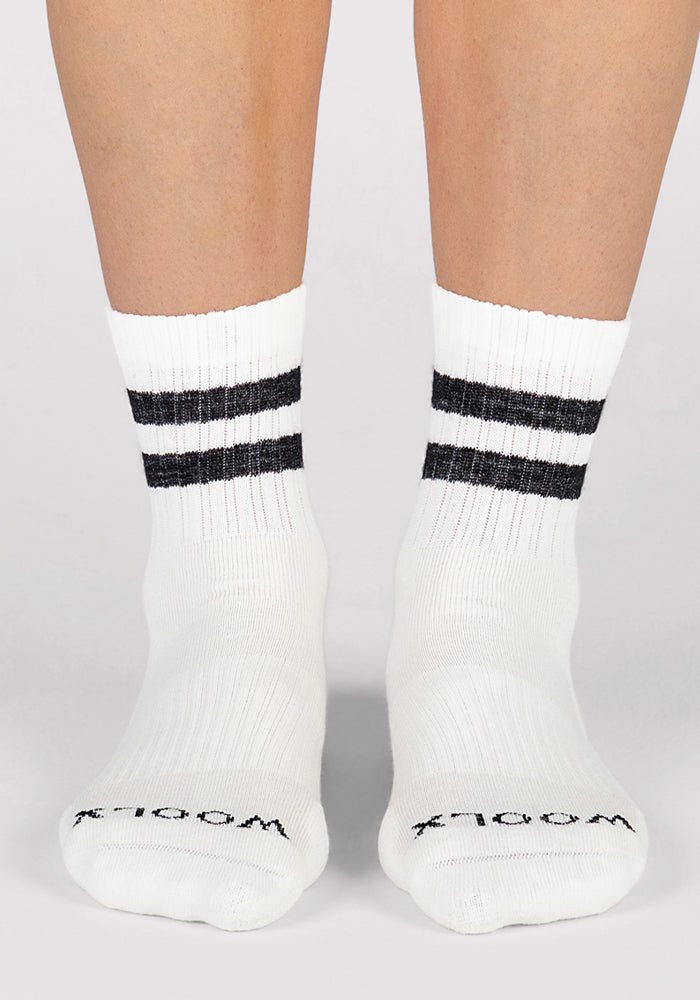 A person wears Woolx Womens Varsity Mini Crew Socks - Classic Stripe, featuring two black stripes at the ankle and "WOOLX" printed in black on the toes. These breathable socks are shown on a plain, light background.