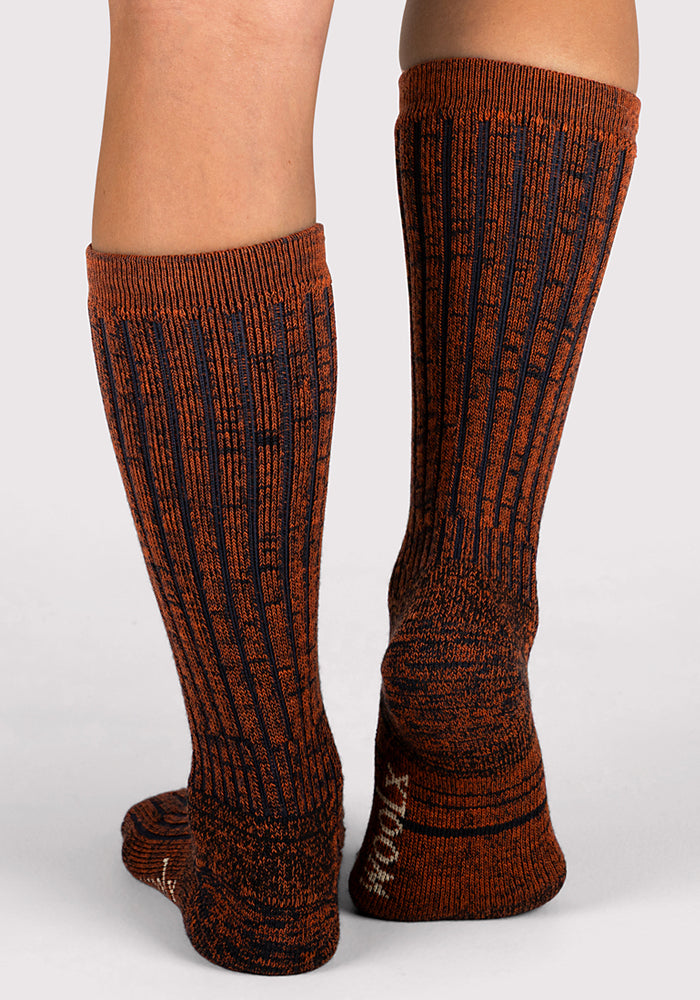 A person wears Woolx Womens Joey Mid Calf Socks in Cinnamon Spice, featuring a tall, ribbed design in burnt orange and black, standing on a light gray surface with feet slightly apart and toes forward.