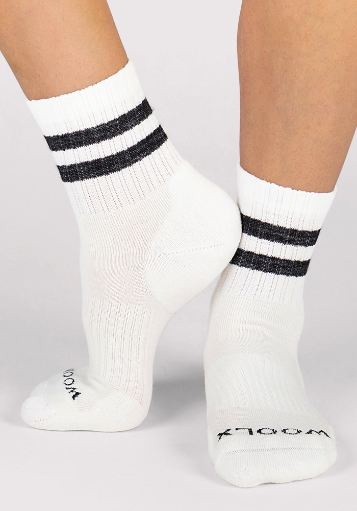 Close-up of two feet in Woolx Womens Varsity Mini Crew Socks - Classic Stripe, featuring white fabric and two black stripes near the top. These mid-ankle socks are breathable and shown on a light background with one heel slightly raised.