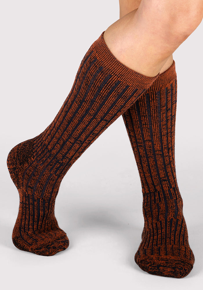 A woman wears Woolx Womens Joey Mid Calf Socks in Cinnamon Spice—a ribbed Merino wool sock with a marled burnt orange and black pattern—standing with one leg crossed before the other against a light gray background.