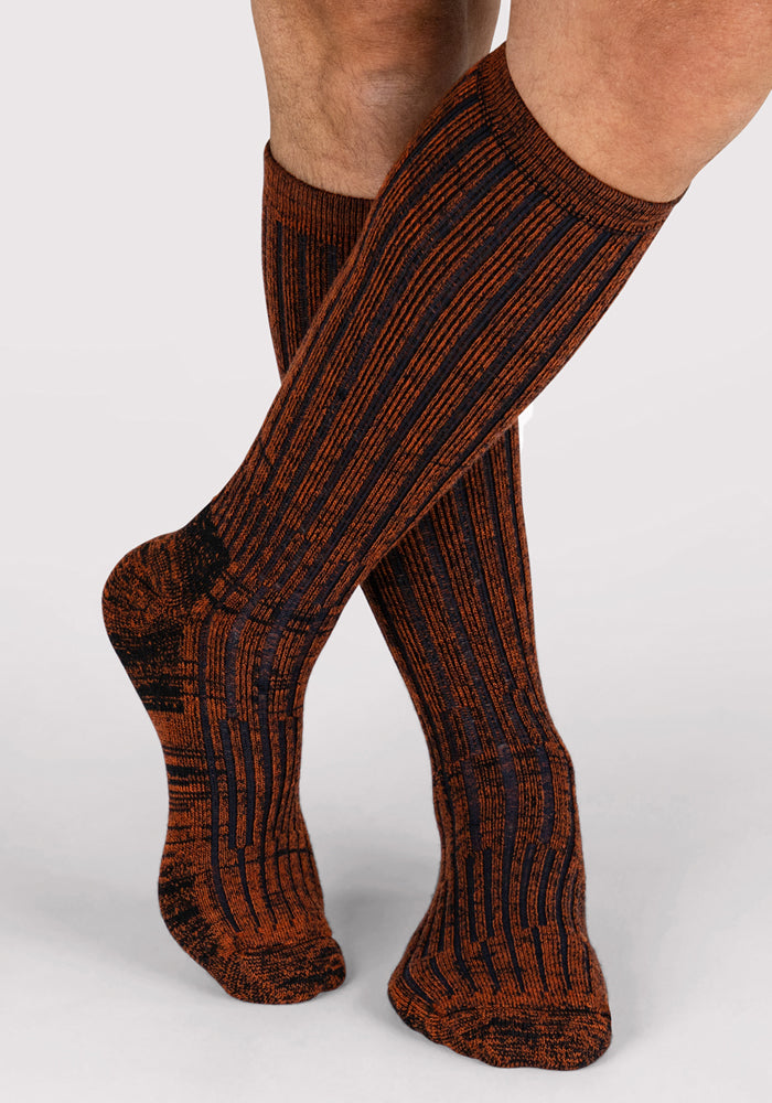 A person wears Woolx Mens Joey Mid Calf Socks in Cinnamon Spice, featuring a brown and black striped pattern. Their moisture-wicking legs are crossed at the ankles against a plain light background.