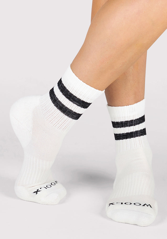 A person crosses their feet at the ankles, wearing Woolx Womens Varsity Mini Crew Socks - Classic Stripe in white with two black stripes near the top and "WOOL" on the toes, against a plain light background.