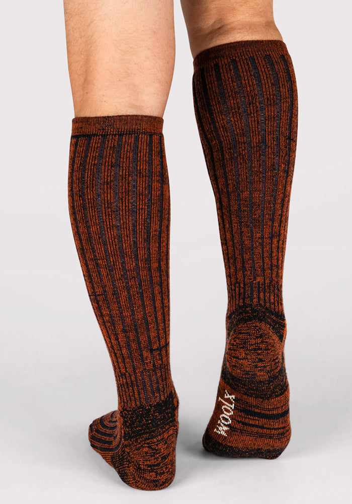 A person wears Woolx Mens Joey Mid Calf Socks in Cinnamon Spice, featuring ribbed orange and black stripes and "WOOL" on the sole, standing on a light gray surface for stylish, moisture-wicking comfort.