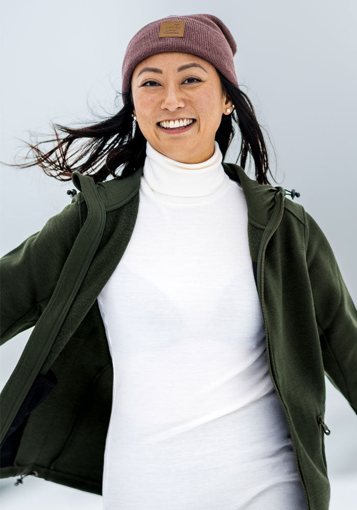 Woman smiles outdoors in Woolx Cubby Hooded Sweatshirt - Duffel Bag, green jacket, and mauve beanie on a snowy day.