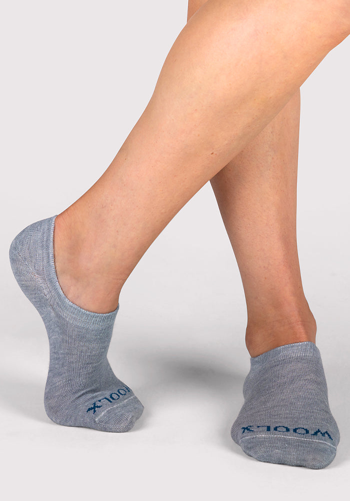 A person wears Woolx Womens Daisy Ped Socks - Calm Waters, light gray merino wool no-show socks with blue text near the toes, standing on a light surface—one foot flat and the other slightly raised—for a soft, stylish women’s look.