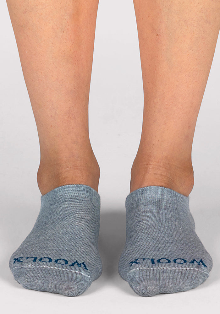 A pair of feet wearing Woolx Womens Daisy Ped Socks in Calm Waters (light gray) with "WOOLX" in blue on the toes stands on a plain white background.