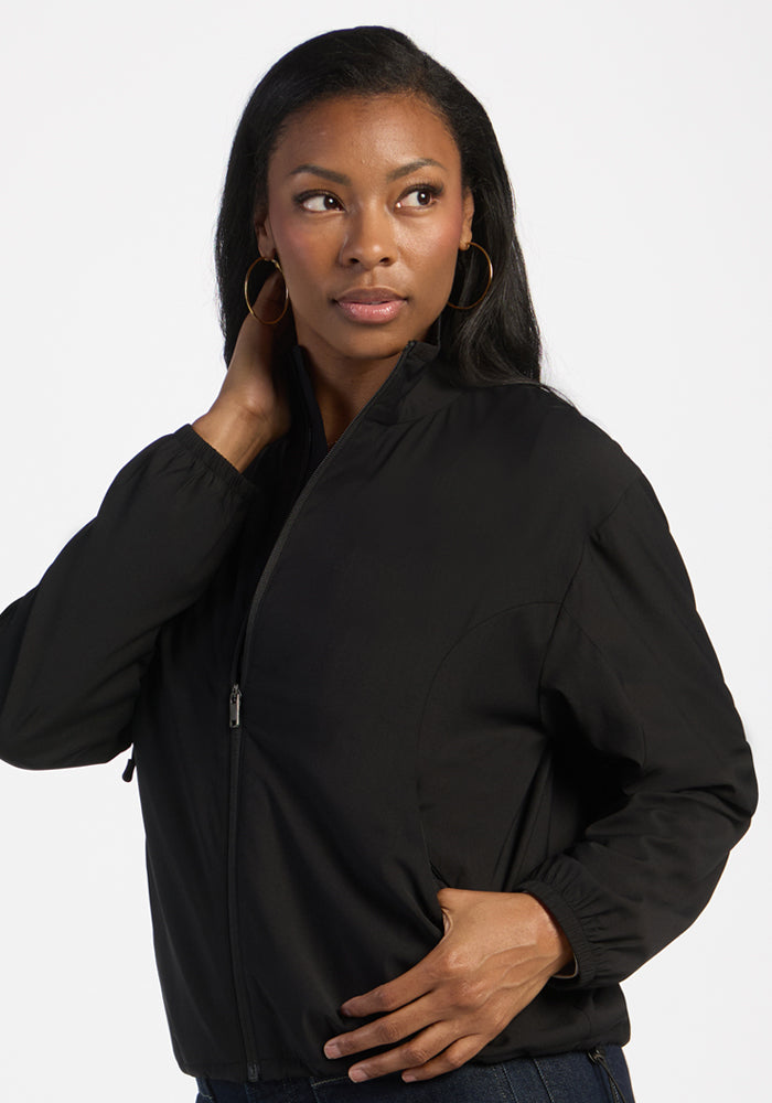 Woman in the Woolx Cooper Windbreaker - Black, zippered pockets, hand on neck, gold hoops, and long dark hair.