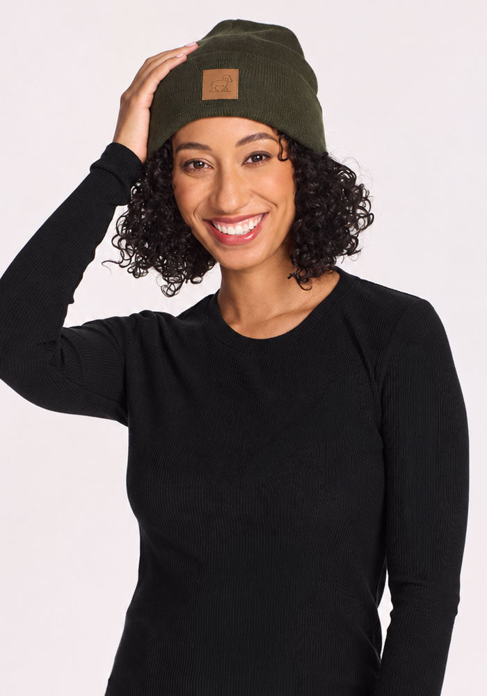 Smiling woman in Woolx Baylor Beanie - Duffel Bag and black top touches her hat against plain light background.