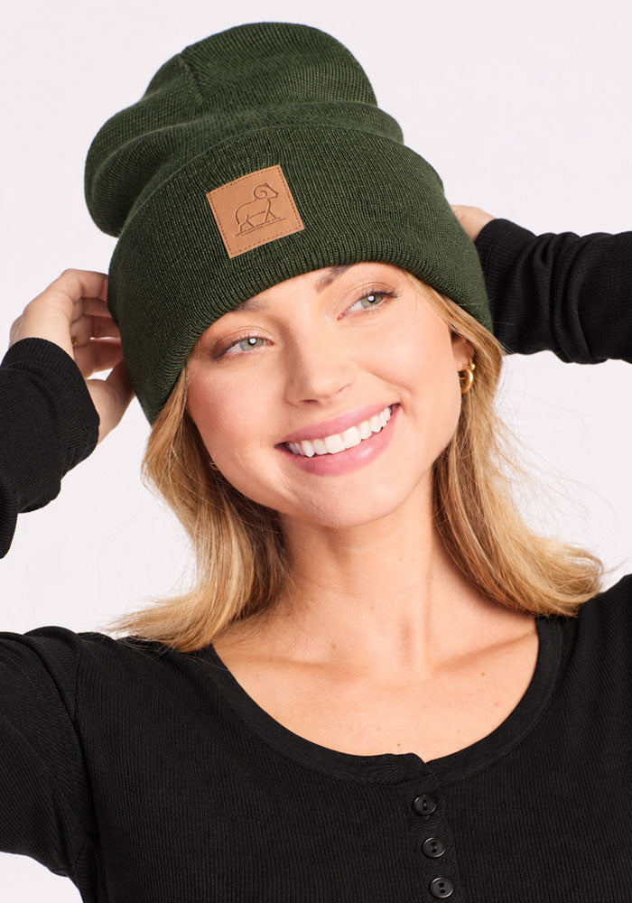 Smiling woman in a Woolx Baylor Beanie - Duffel Bag and black shirt, holding her warm winter hat with both hands.