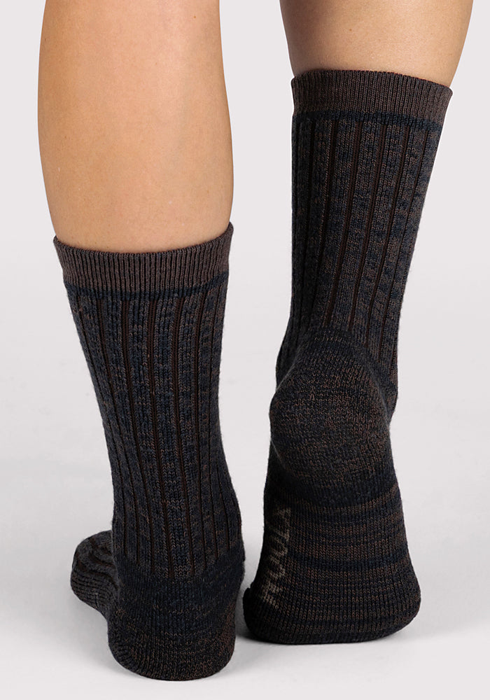 A close-up shows the back of a person's lower legs and feet wearing Woolx Womens Joey Crew Socks in Dark Espresso, standing on a light neutral background.