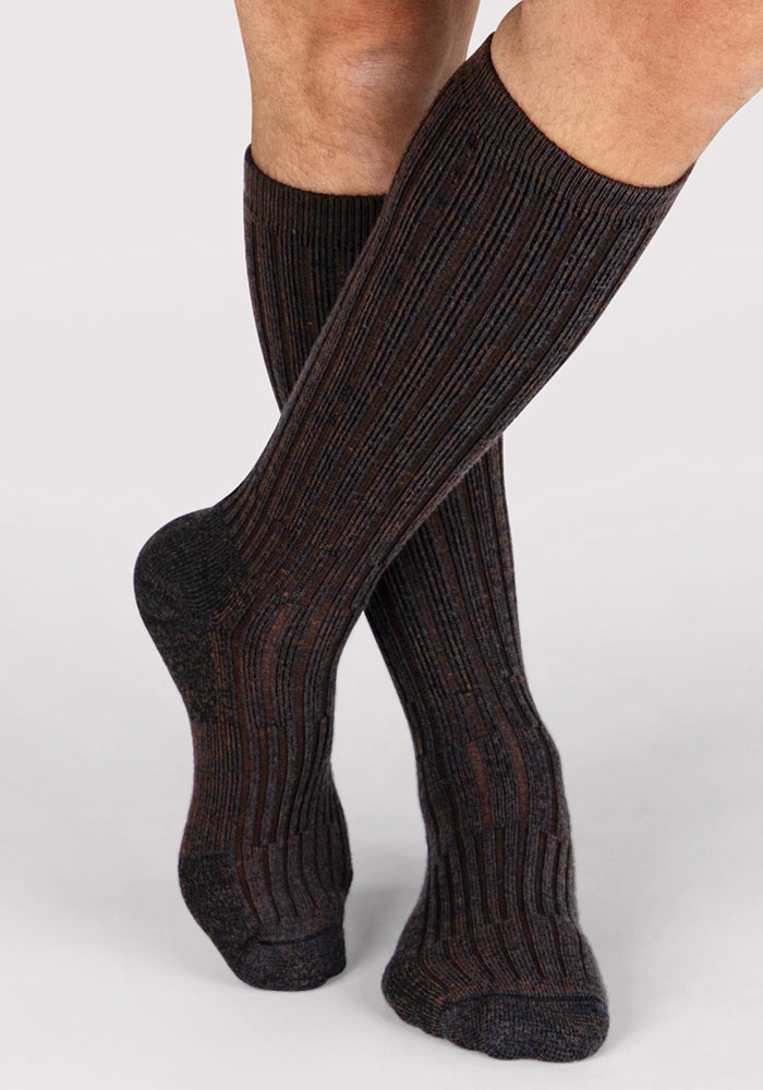 A person stands on tiptoe wearing Woolx Mens Joey Mid Calf Socks in Dark Espresso, featuring ribbed merino wool with subtle vertical stripes. The background is plain and light gray.