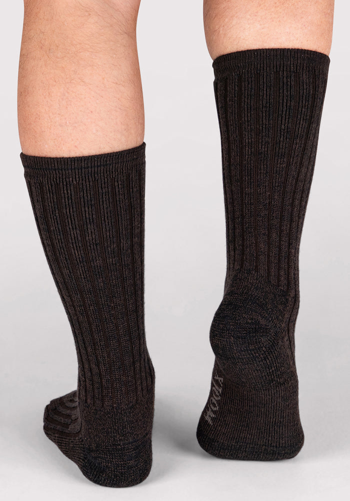 A person stands on a light surface wearing Woolx Mens Joey Crew Socks in Dark Espresso. Only their lower legs and feet are visible, with one foot slightly raised at the heel.