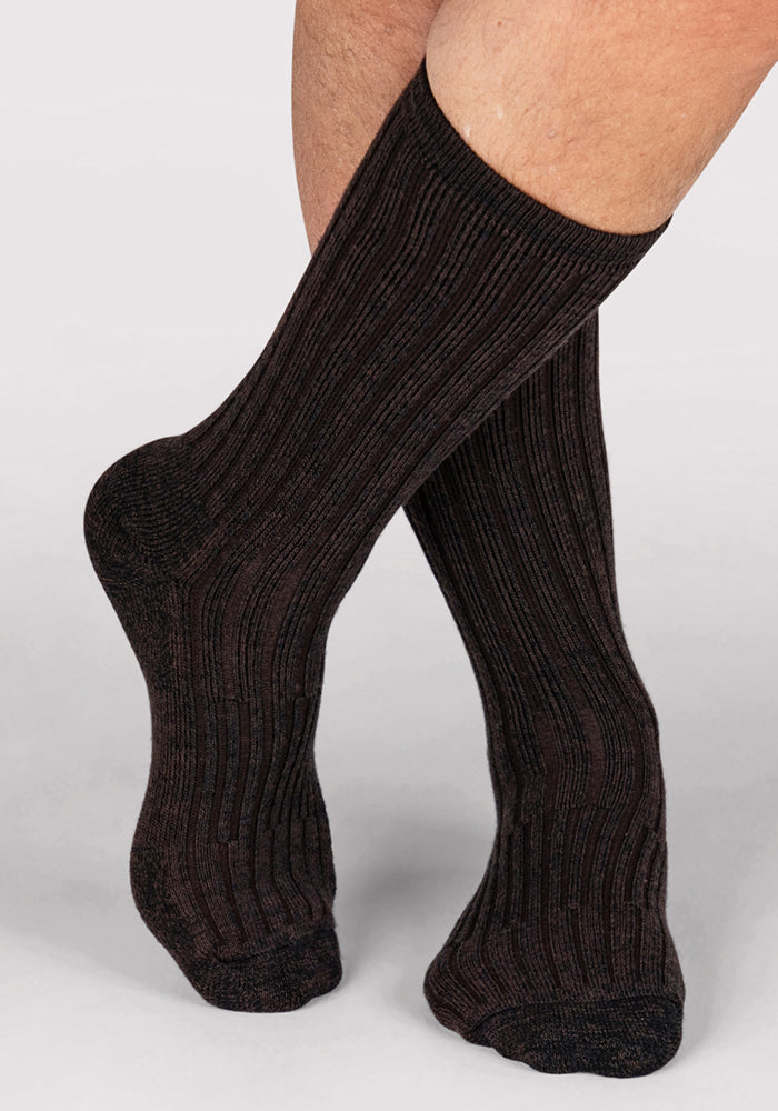 A close-up of a person's lower legs wearing Woolx Mens Joey Crew Socks in Dark Espresso, standing on a light gray surface with one foot slightly ahead of the other.