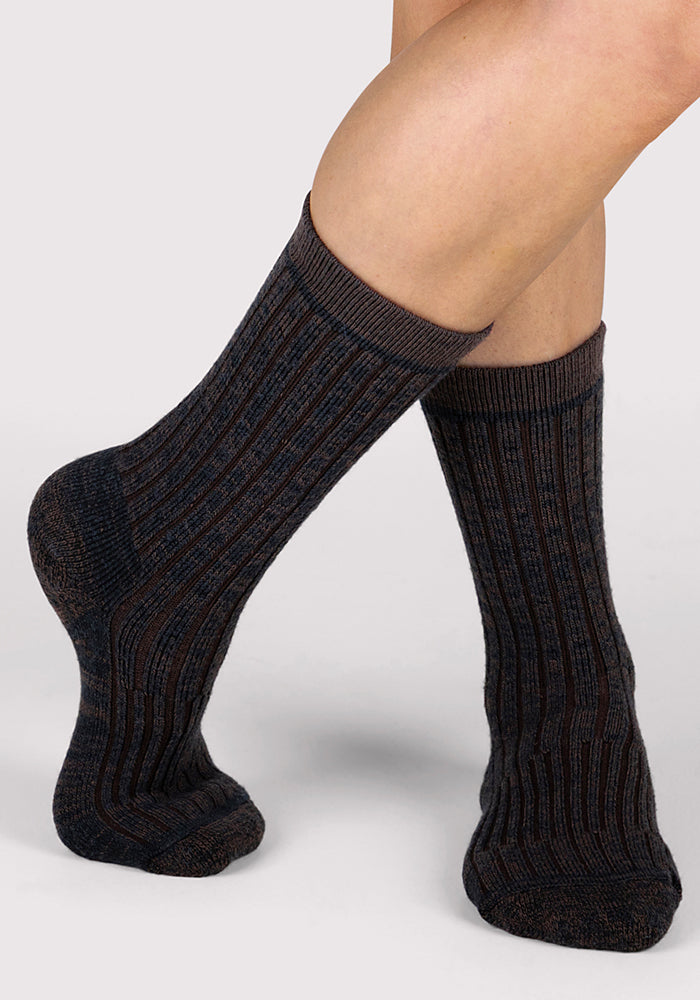 A person wearing Woolx Womens Joey Crew Socks in Dark Espresso stands with one foot slightly raised and crossed over the other against a light gray background.