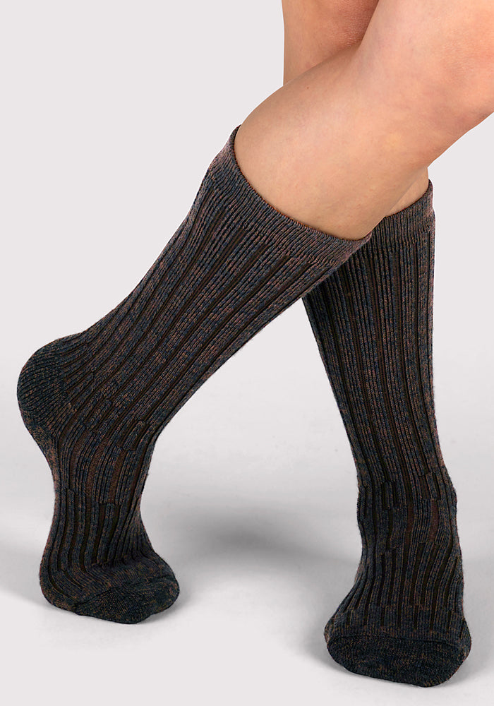 A person with bare legs is wearing Woolx Womens Joey Mid Calf Socks in Dark Espresso, which are thick and ribbed. The socks reach just below the knees, feet are crossed at the ankles, and the background is plain light gray.