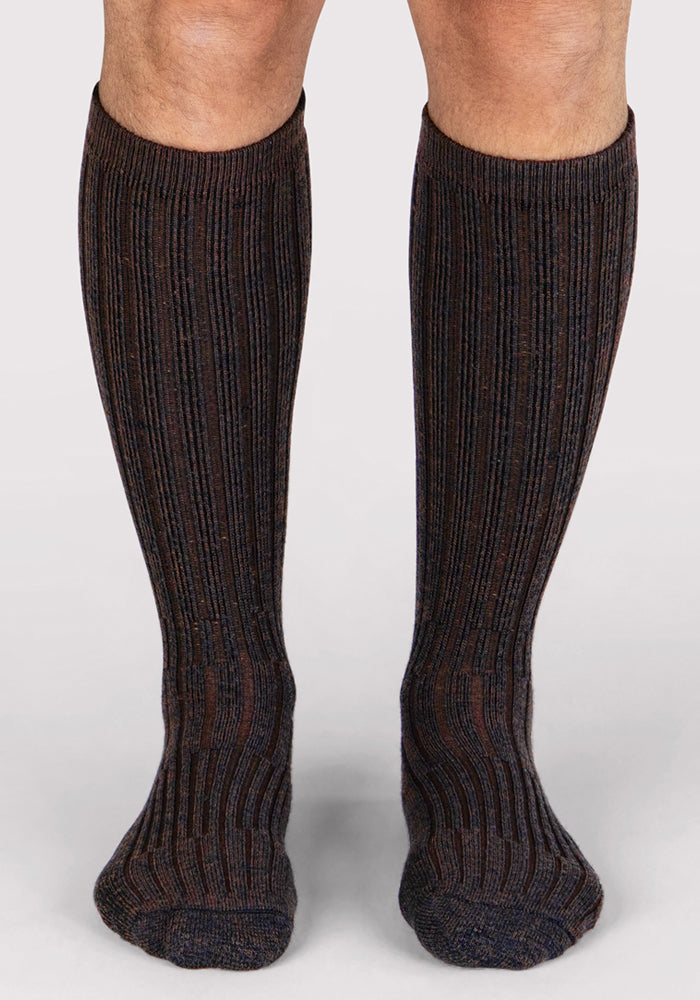 A person stands on a light background wearing Woolx Mens Joey Mid Calf Socks in Dark Espresso, featuring a ribbed design and subtle stripes crafted from warm Merino wool.