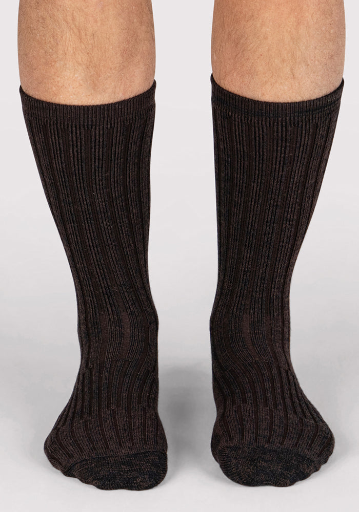 A close-up of a person's lower legs showcases the Woolx Mens Joey Crew Socks in Dark Espresso, featuring a ribbed design, as they stand on a light gray surface.
