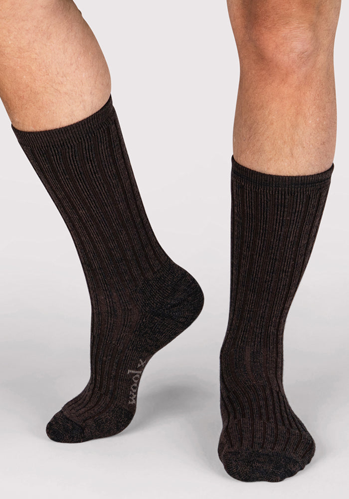 A person is wearing Woolx Mens Joey Crew Socks in Dark Espresso, standing on a light-colored surface. Only their lower legs and feet are visible.