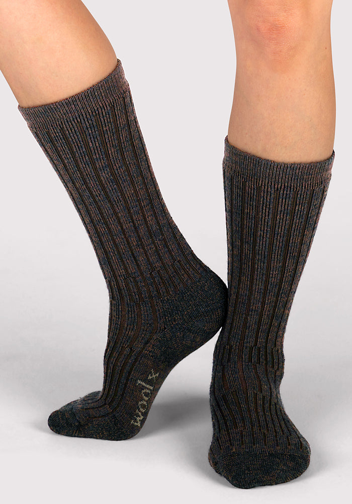 A person wearing Woolx Womens Joey Mid Calf Socks in Dark Espresso, featuring ribbed fabric and a subtle striped pattern, stands on tiptoe with one leg bent and the brand name visible on the sole.