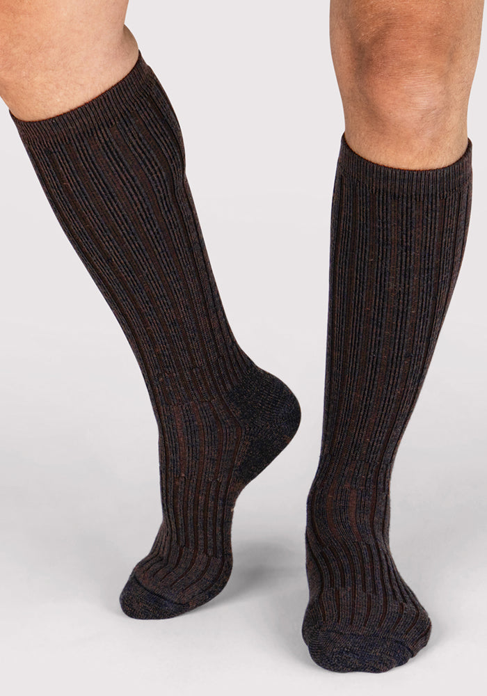 A person wearing Woolx Mens Joey Mid Calf Socks in Dark Espresso stands on a light-colored surface, one foot slightly ahead of the other.