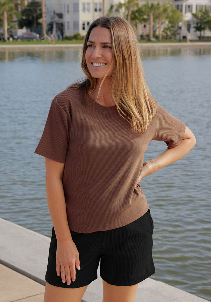 A woman with long brown hair smiles by the waterfront, wearing the Woolx Delia Ribbed Short Sleeve in Sienna Kiss and black shorts. Trees and buildings are visible across the water in the background.