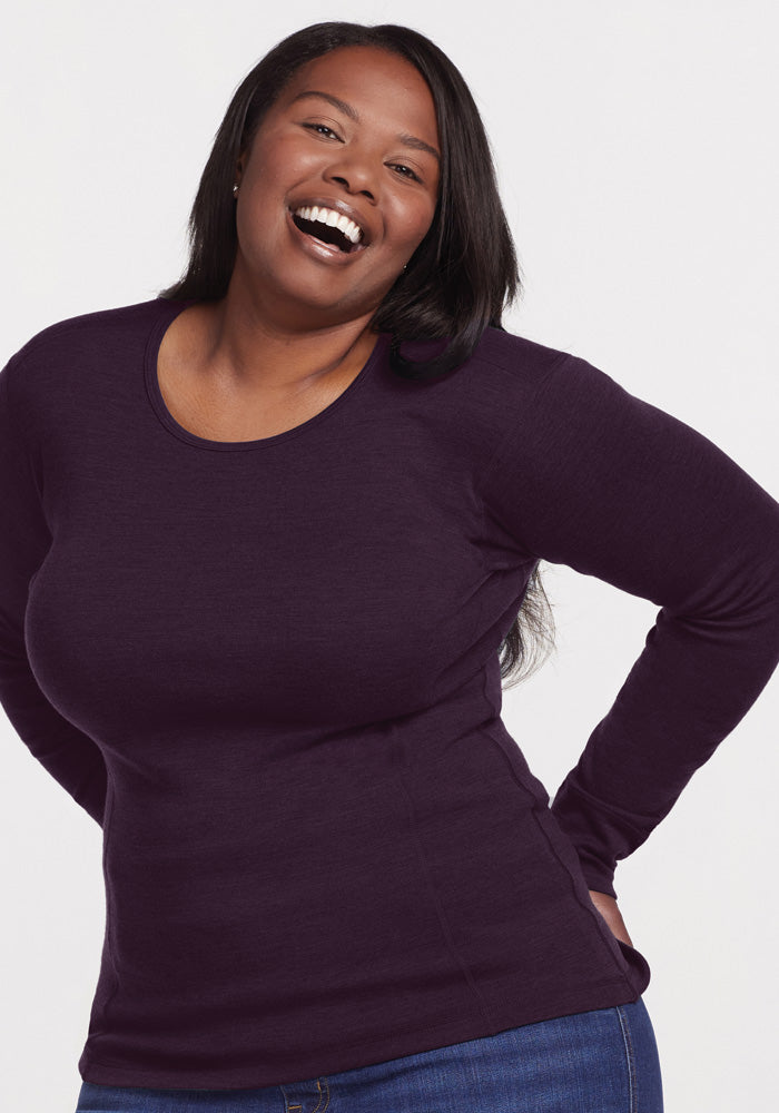 A woman smiles, wearing the Woolx Hannah Long Sleeve Top in Deep Plum paired with blue jeans, standing with hands on hips against a white backdrop.