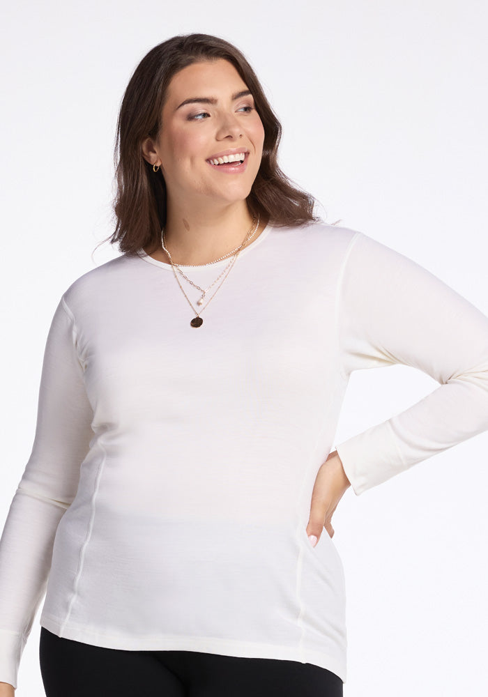 A woman beams confidently to the side, standing against a plain white background. She dons layered necklaces and black pants, with her hand on her hip, showcasing the moisture-wicking Hannah Long Sleeve Top in Buttercream by Woolx.