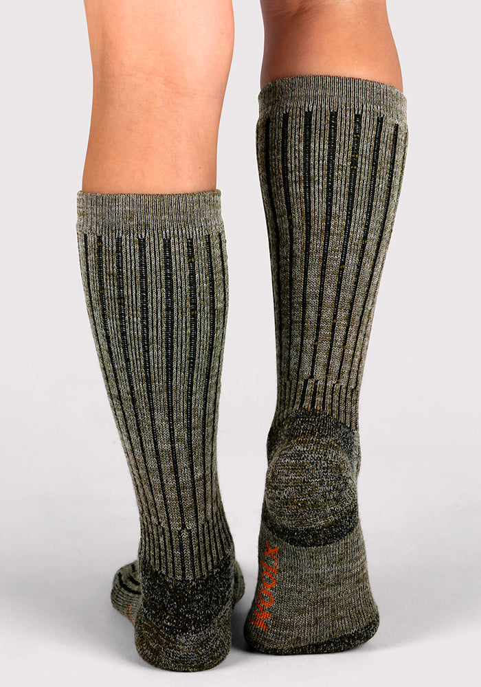 A person stands on a light background, showing the back of their lower legs and feet while wearing Woolx Womens Joey Mid Calf Socks in Dark Forest Marled.