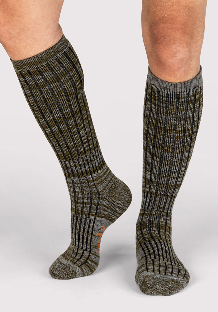 A person stands on a light background, one foot forward, wearing Woolx Mens Joey Mid Calf Socks in Dark Forest Marled—ribbed merino wool socks in olive green, brown, and gray.