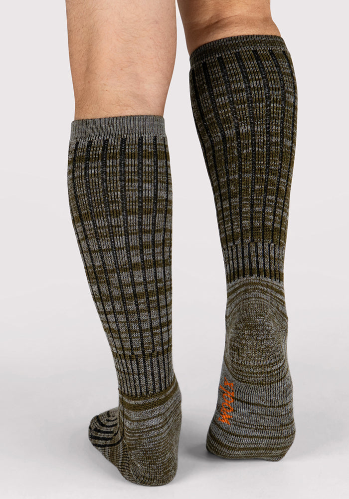 A person stands on a light gray surface wearing Woolx Mens Joey Mid Calf Socks in Dark Forest Marled, featuring a ribbed texture in gray and olive green with an orange logo on the sole.