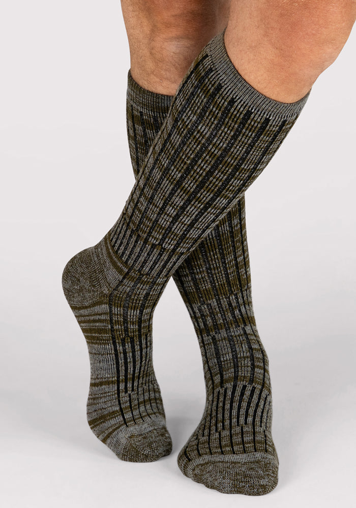 A person wearing Woolx Mens Joey Mid Calf Socks in Dark Forest Marled stands with legs crossed at the ankles against a plain light background.