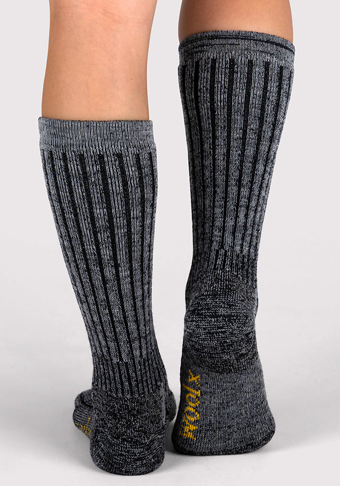A close-up of two legs wearing Woolx Womens Joey Mid Calf Socks in dark navy marled, featuring ribbed Merino wool, black stripes, and yellow writing on the heels. The background is plain and light-colored.